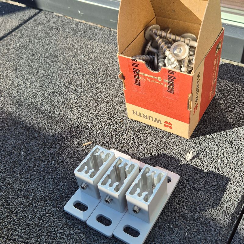 Tre braketter i aluminium lagt klar ved siden av en eske skruer